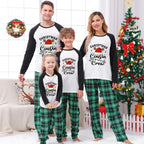 A family of four wearing matching Christmas-themed raglan pajamas stands together in a decorated living room with a Christmas tree, wreath, and holiday decor. The pajama set has a white front with black sleeves and a printed graphic reading 'Christmas Cousin Crew' with a Santa hat, plus green plaid pants.
