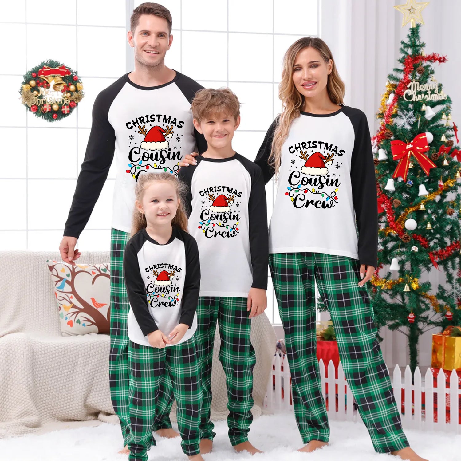 A family of four wearing matching Christmas-themed raglan pajamas stands together in a decorated living room with a Christmas tree, wreath, and holiday decor. The pajama set has a white front with black sleeves and a printed graphic reading 'Christmas Cousin Crew' with a Santa hat, plus green plaid pants.