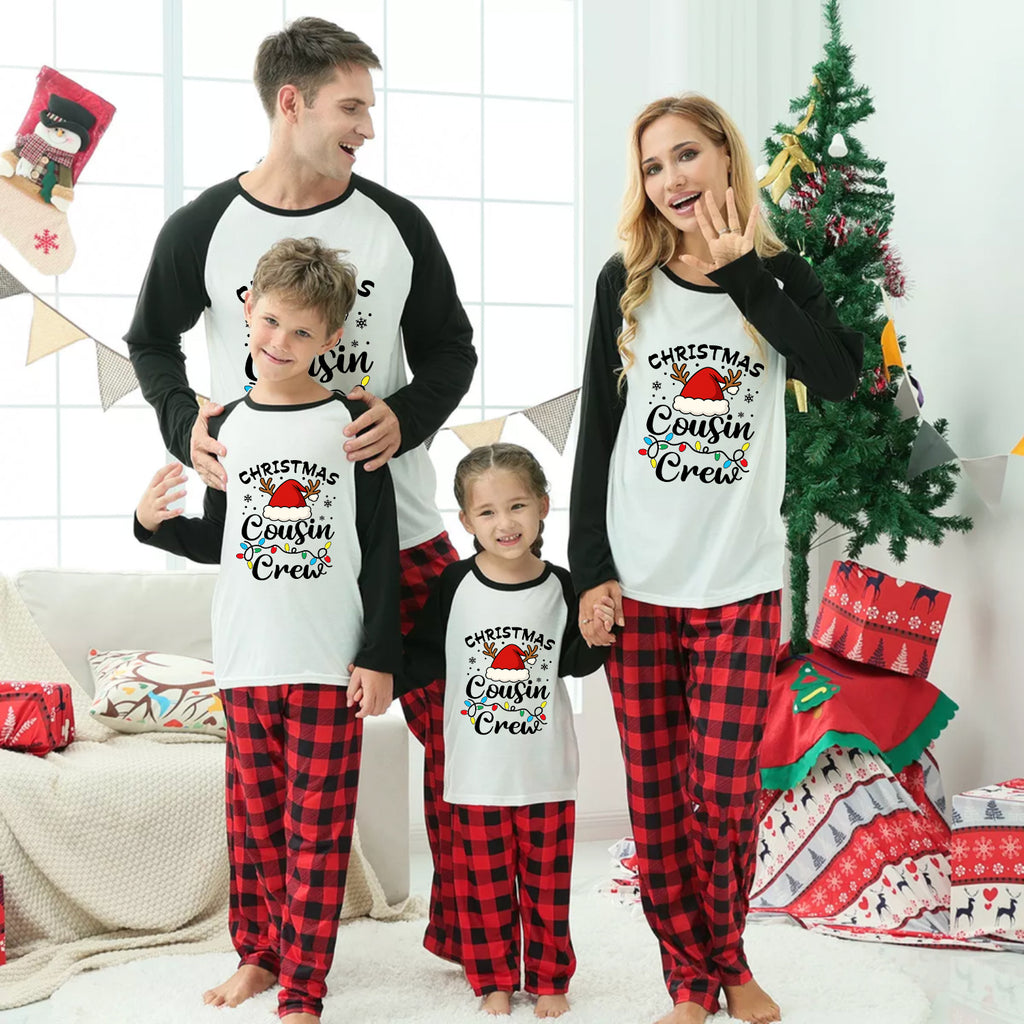 A family of four wearing matching Christmas-themed pajamas stands together in a living room decorated for Christmas. The matching set features a white and black long-sleeve top with 'CHRISTMAS Cousin Crew' text and a red and black buffalo plaid pant leg.