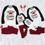 A family Christmas cousin crew pajama set laid out on a white textured background, including four raglan long-sleeve shirts in different sizes with black sleeves and black collar trim, printed with 'Christmas Cousin Crew' and a Santa hat graphic, matching red and black plaid pants, a red snowflake headband, and a red snowflake accessory.