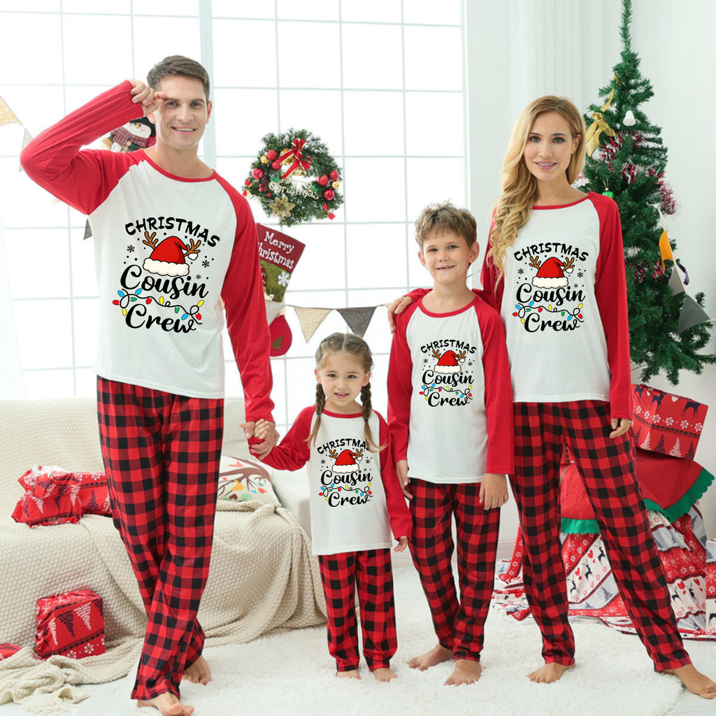 A family of four wearing matching Christmas-themed long-sleeve pajamas stands together in a decorated living room. The pajamas have red and black buffalo plaid legs with white fronts and red sleeves, featuring a festive Santa graphic.