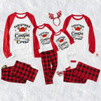 A family matching Christmas cousin crew pajama set laid out on a white textured background, including long-sleeve raglan shirts and plaid pants in red and black, plus a red reindeer antler headband.