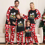 A family of four wearing matching holiday pajamas standing in a living room. The pajamas feature a black front with a colorful gnome graphic and red-and-black buffalo plaid sleeves and pants.
