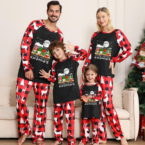 A family of four wearing matching holiday pajamas standing in a living room. The pajamas feature a black front with a colorful gnome graphic and red-and-black buffalo plaid sleeves and pants.