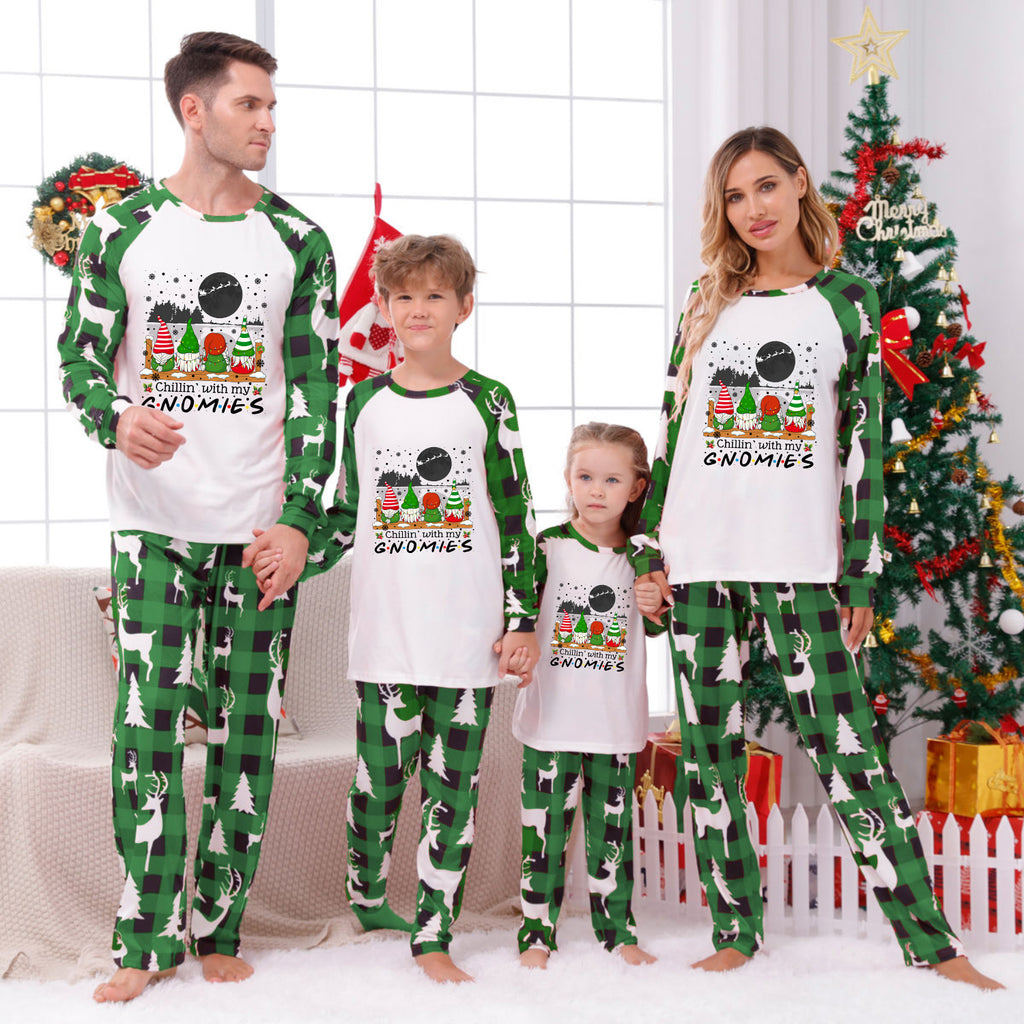 A family of four wearing matching Christmas-themed pajamas, standing in a living room decorated for Christmas. The pajamas feature a white front with a colorful gnome graphic and green raglan sleeves and pants with a green and black gingham pattern.
