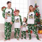 A family of four wearing matching Christmas-themed pajamas, standing in a living room decorated for Christmas. The pajamas feature a white front with a colorful gnome graphic and green raglan sleeves and pants with a green and black gingham pattern.