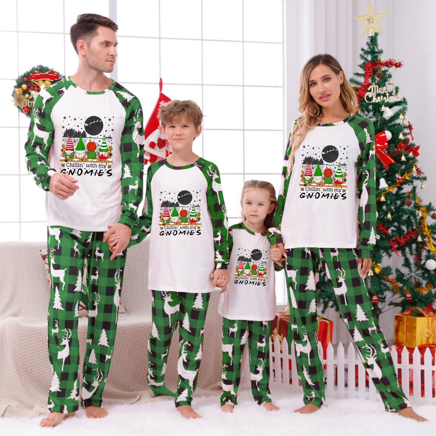 A family of four wearing matching Christmas-themed pajamas, standing in a living room decorated for Christmas. The pajamas feature a white front with a colorful gnome graphic and green raglan sleeves and pants with a green and black gingham pattern.