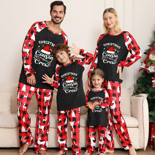 A family of four wearing matching Christmas-themed long-sleeve pajamas, standing together in front of a beige couch with a decorated Christmas tree on the right. The pajamas feature a black front with white text reading 'Christmas Cousin/Crew' and a red-and-white buffalo plaid pattern on the sleeves and pants, with white reindeer prints.