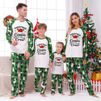 Three matching family Christmas pajama sets in different sizes, featuring cream-colored long-sleeve tops with a black-and-white striped Christmas tree graphic and patterned pants. The pants have holly and pinecone motifs, with the largest set having dark brown pinecones and the smaller sets having dark brown pinecones with green holly accents.