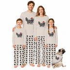 A family of four wearing matching Christmas-themed pajamas stands together on a white background, with a small dog sitting at their right. The pajamas are light beige long-sleeve tops with a colorful raccoon graphic wearing a Santa hat, paired with black-and-white cat-print pants.