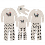 A matching family pajama set displayed on a white background, including three sizes of long-sleeve shirts and pants. The shirts are cream-colored with a raccoon graphic decorated with colorful Christmas lights and a red Santa hat.
