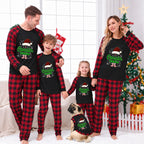 A family of four and a dog wearing matching Christmas pajamas in a living room setting with a decorated Christmas tree. The pajamas feature a black raglan top and red-and-black buffalo plaid pants or matching top-and-pants sets, with a 'Cousin Crew' graphic on the front.