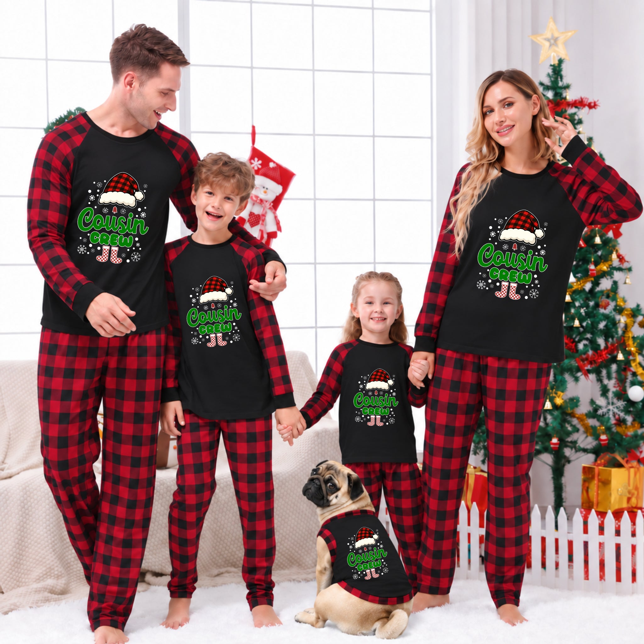 A family of four and a dog wearing matching Christmas pajamas in a living room setting with a decorated Christmas tree. The pajamas feature a black raglan top and red-and-black buffalo plaid pants or matching top-and-pants sets, with a 'Cousin Crew' graphic on the front.