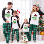 A family of four and a dog wearing matching Christmas raglan pajamas in a decorated living room. The matching set features white long-sleeve tops with black sleeves and green plaid pants