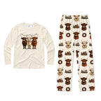 A two-piece pajama set on a White background, featuring a Cream long-sleeve shirt with a graphic of three Highland cattle under Christmas lights, and matching Cream pants with an all-over print of Highland cattle, wreaths, and snowflakes.