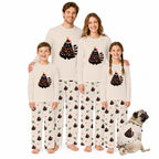 A family of five wearing matching Christmas-themed pajamas stands together on a white background, with a small dog sitting at their right. The pajamas are cream-colored with a black tree graphic decorated with red ornaments, yellow lights, and a green Santa hat, paired with matching patterned pants.