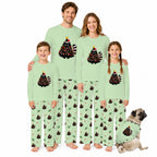 A family of five wearing matching light green Christmas-themed pajamas stands together on a white background, with a small pug dog wearing a matching pajama set beside them. The pajamas feature a black Christmas tree graphic with red ornaments and a striped tail on the chest, and a repeating pattern of dark pine trees with small red and green accents on the pants.