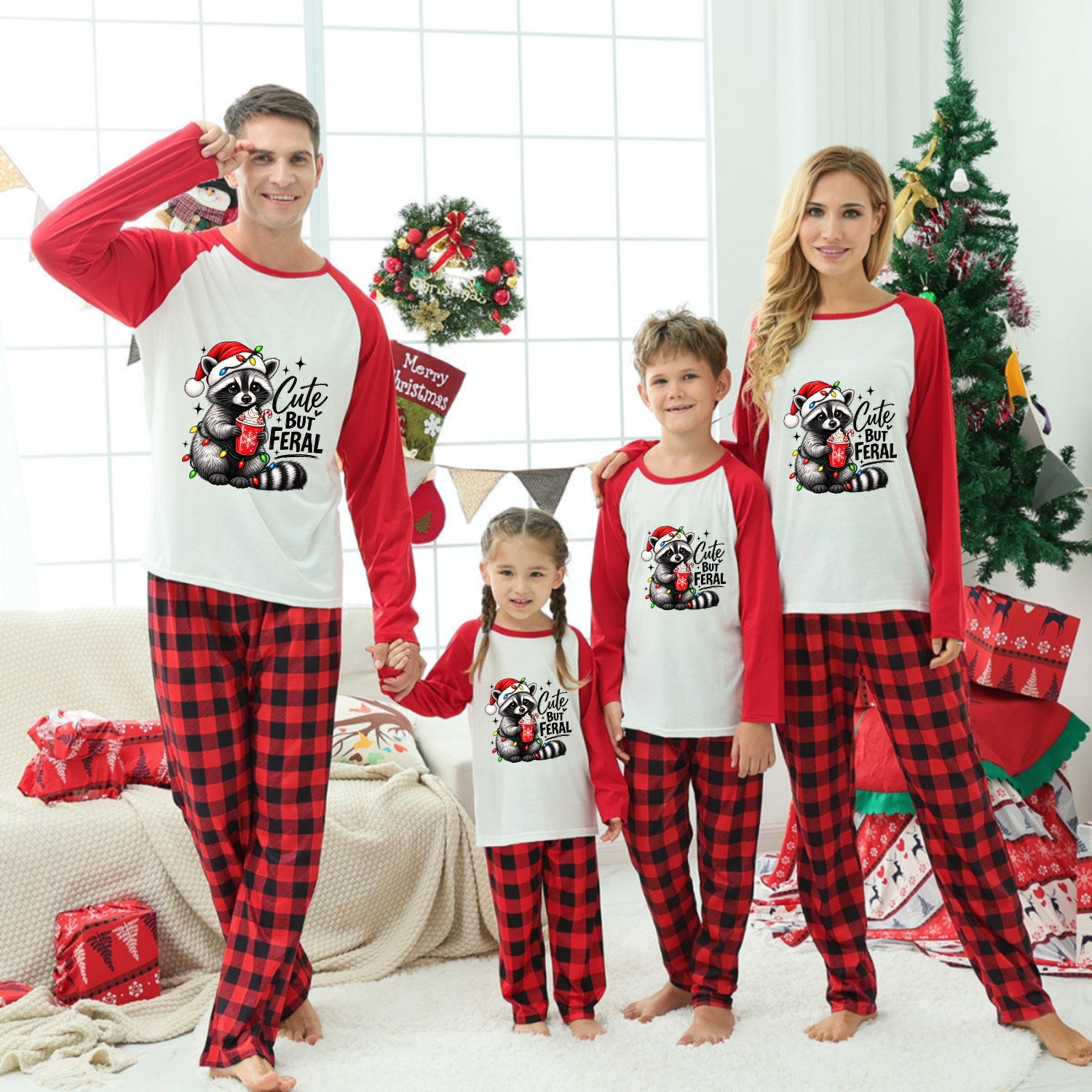 A family of four wearing matching Christmas-themed long-sleeve pajamas stands together in a decorated living room. The pajamas have red and black buffalo plaid legs with white fronts and red sleeves, featuring a festive Santa graphic.