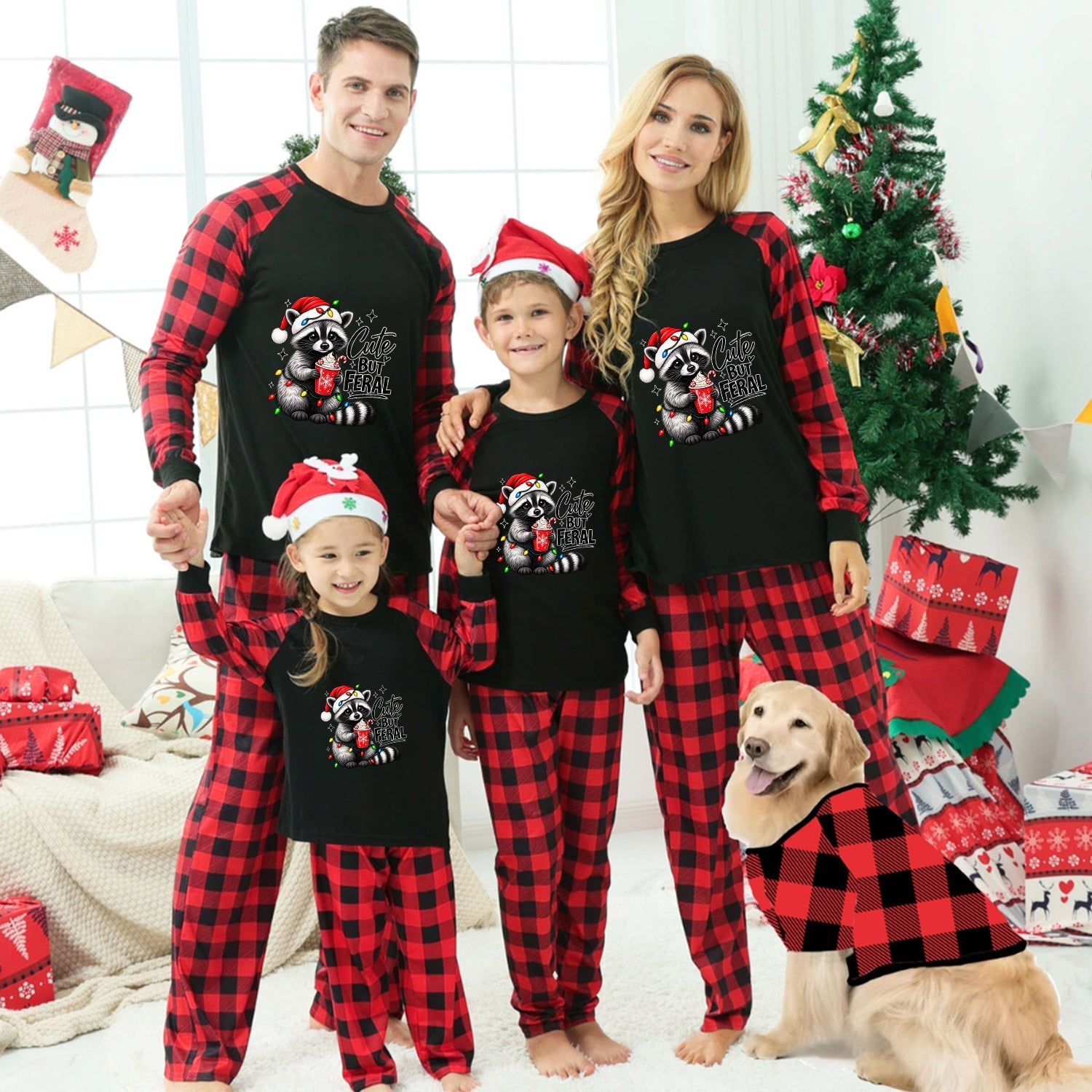 A family of four wearing matching black and red plaid Christmas pajamas with a raccoon graphic, standing in a decorated living room with a dog wearing a matching pajama set. The pajamas feature a black body with red and black buffalo plaid sleeves and legs, and the front has a graphic of a raccoon holding a Christmas tree with the text 'Cute but Feral'.