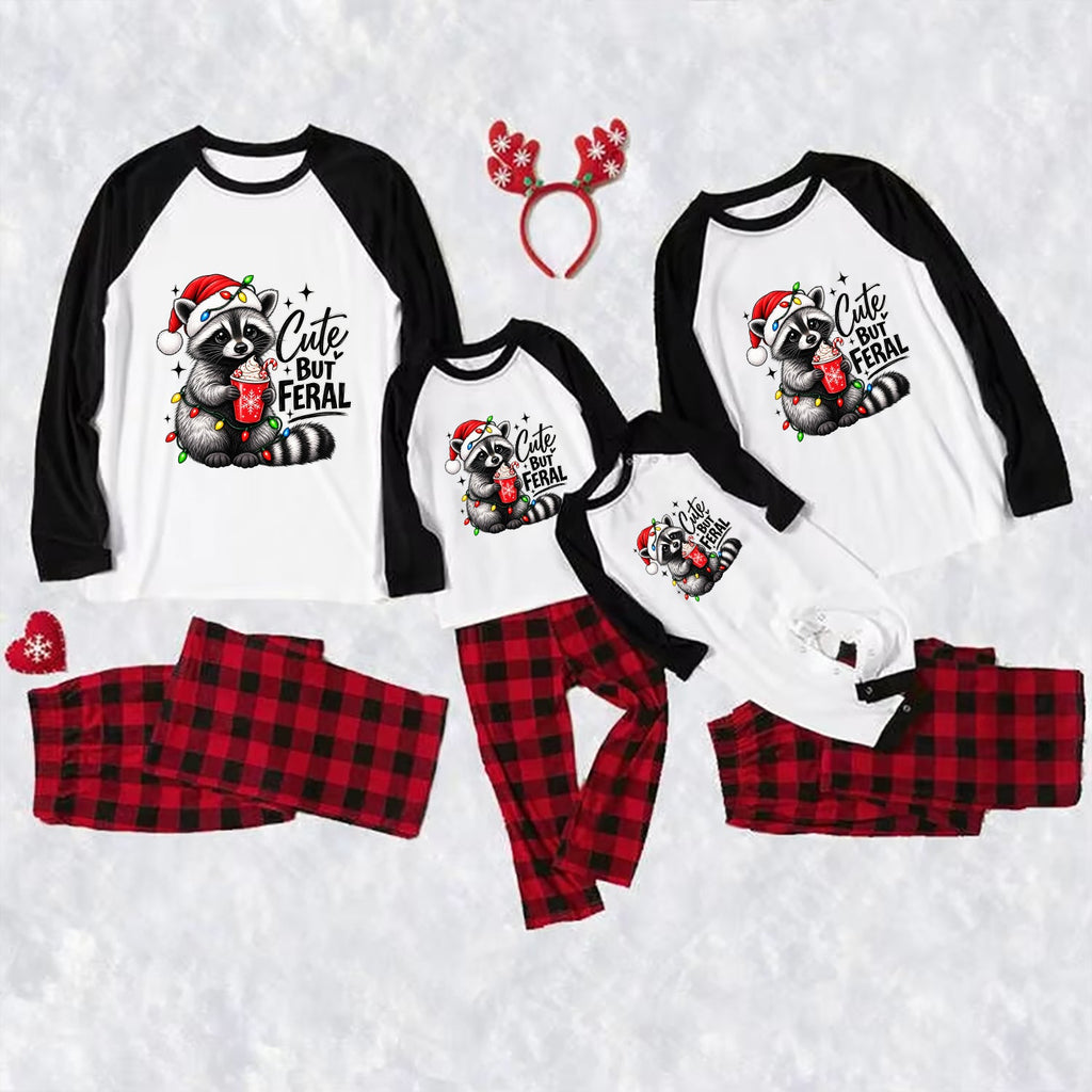 Three matching family Christmas raglan pajamas laid out on a white textured background, including large, medium, and small long-sleeve shirts with black sleeves and a raccoon graphic that reads 'Cute But Feral,' plus red-and-black plaid pants. A red reindeer antler headband and small red heart accessory are also included.