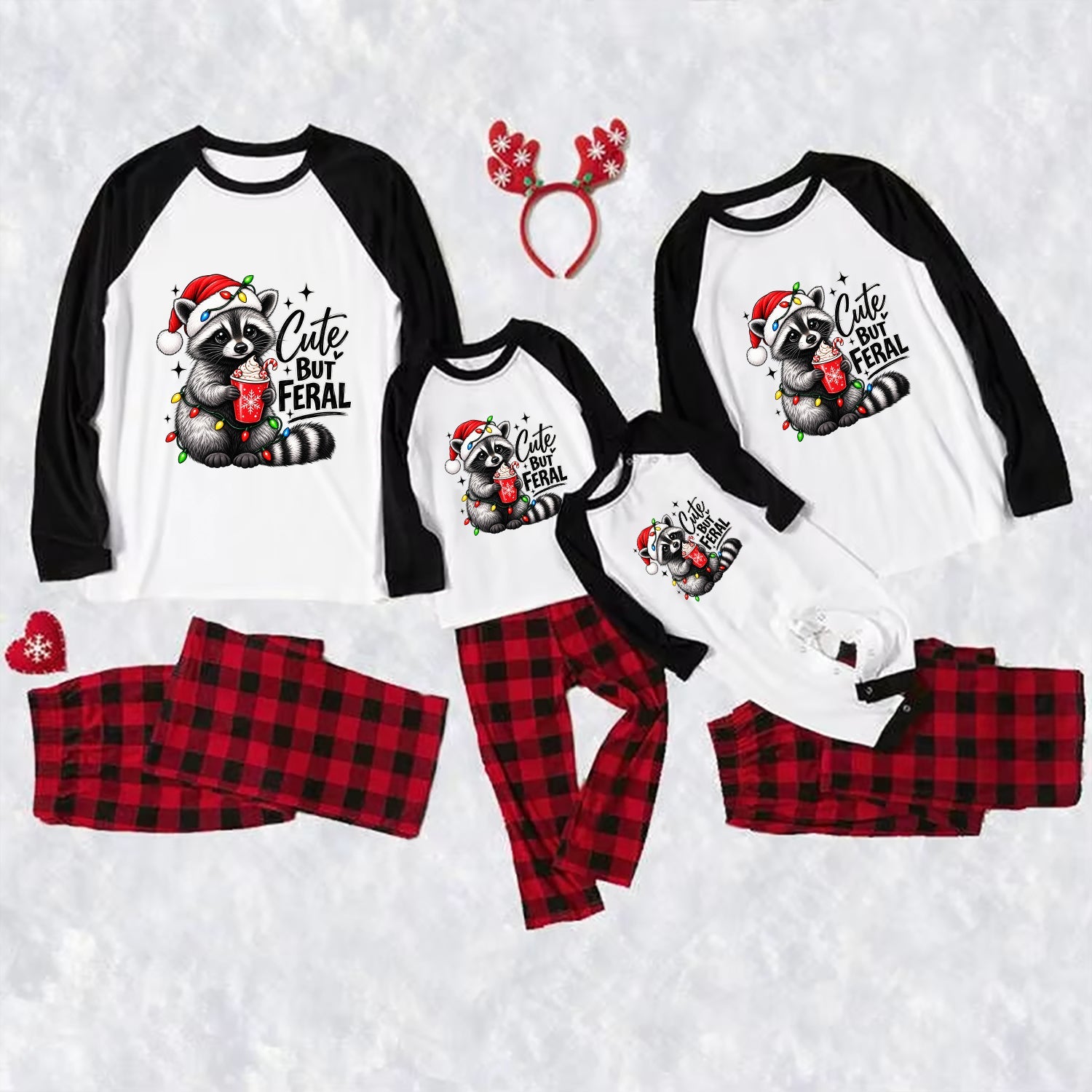 Three matching family Christmas raglan pajamas laid out on a white textured background, including large, medium, and small long-sleeve shirts with black sleeves and a raccoon graphic that reads 'Cute But Feral,' plus red-and-black plaid pants. A red reindeer antler headband and small red heart accessory are also included.