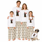 A family of four and a dog wearing matching Christmas pajamas against a plain White background. The pajamas are White long-sleeve tops with a colorful dog graphic wearing a Santa hat, paiRed with Beige pants coveRed in a repeating dog pattern.
