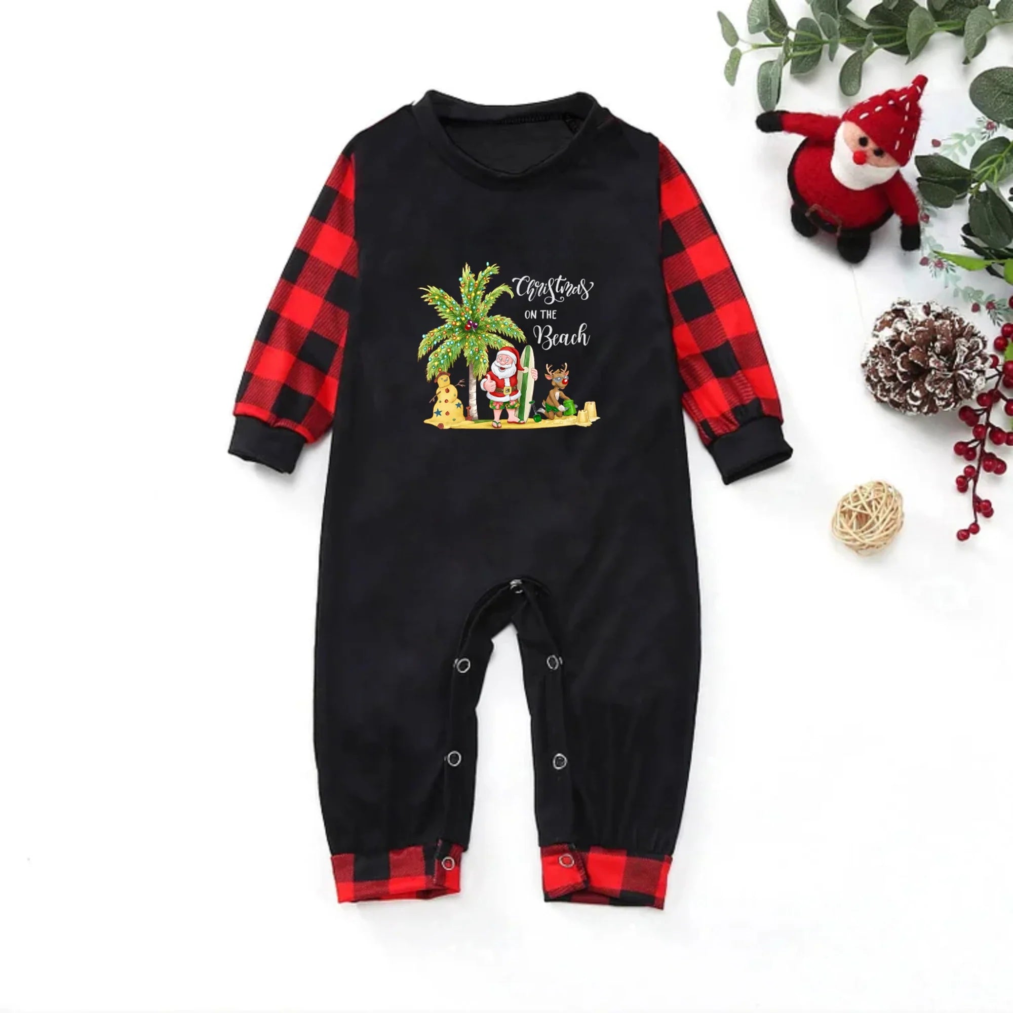 A baby long-sleeve romper in Black with Red buffalo plaid sleeves and cuffs, featuring a Christmas-themed graphic on the chest with a palm tree, Santa, and the text 'Christmas is Coming'. The romper is displayed flat on a White background with holiday props like a small Santa doll, pinecones, and Greenery.