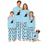 A family of five wearing matching light blue Christmas-themed pajamas with a black cat wearing a Santa hat and green/yellow holiday accents, standing against a white background with a small matching dog at their side.