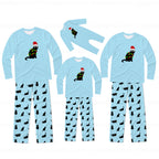 Three sizes of matching family pajama sets shown on a white background. Each set includes a light blue long-sleeve top with a black cat wearing a Christmas tree and Santa hat graphic, paired with light blue pants covered in a black cat print.