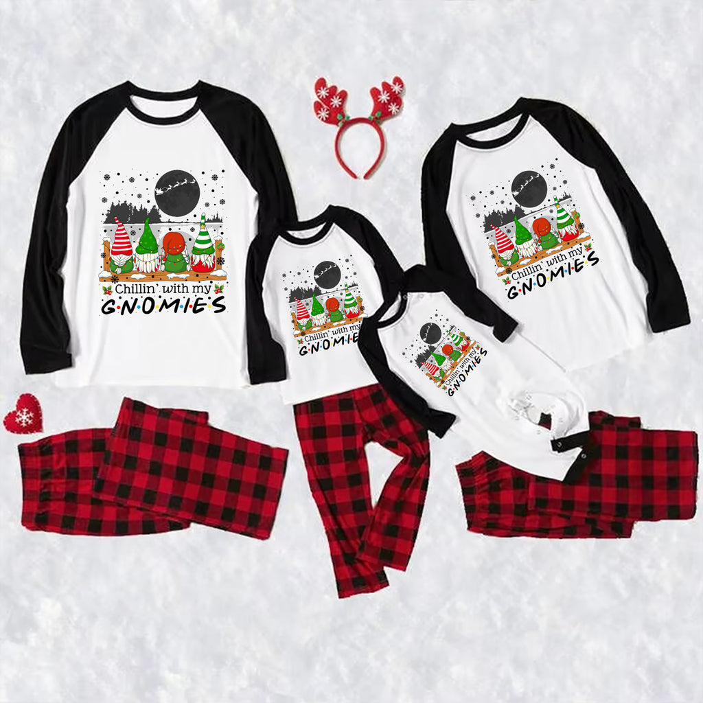 A matching family holiday pajama set laid out on a white textured background, including four raglan long-sleeve shirts with black sleeves and a gnomes graphic on the front, matching red-and-black plaid pants, a red reindeer antler headband, and a small red heart-shaped accessory.