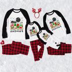 A matching family holiday pajama set laid out on a white textured background, including four raglan long-sleeve shirts with black sleeves and a gnomes graphic on the front, matching red-and-black plaid pants, a red reindeer antler headband, and a small red heart-shaped accessory.