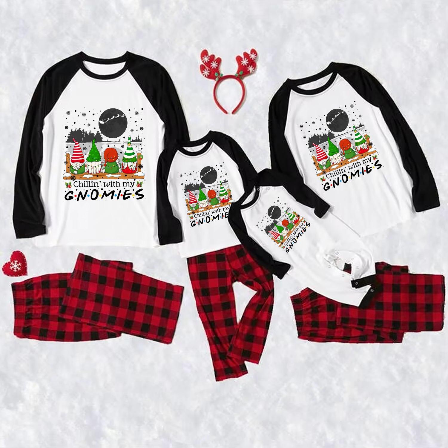 A matching family holiday pajama set laid out on a white textured background, including four raglan long-sleeve shirts with black sleeves and a gnomes graphic on the front, matching red-and-black plaid pants, a red reindeer antler headband, and a small red heart-shaped accessory.