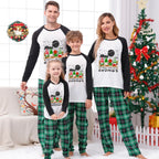 A family of five wearing matching Christmas-themed pajamas stands together on a white background, with a small dog sitting at their right. The pajamas are cream-colored with a black tree graphic decorated with red ornaments, yellow lights, and a green Santa hat, paired with matching patterned pants.