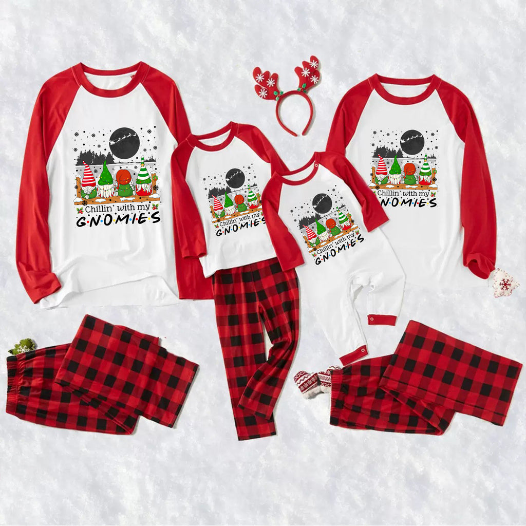A family matching holiday pajama set laid out on a white textured background, including three long-sleeve raglan tops with red sleeves and red gnomes graphics, three matching pants in red buffalo plaid, a red reindeer antler headband, and small red-and-white booties. The tops read 'Cabin with my GNOMIES' and the pants say 'ALL IN A RED AND BLACK BUFFALO PLaid'.