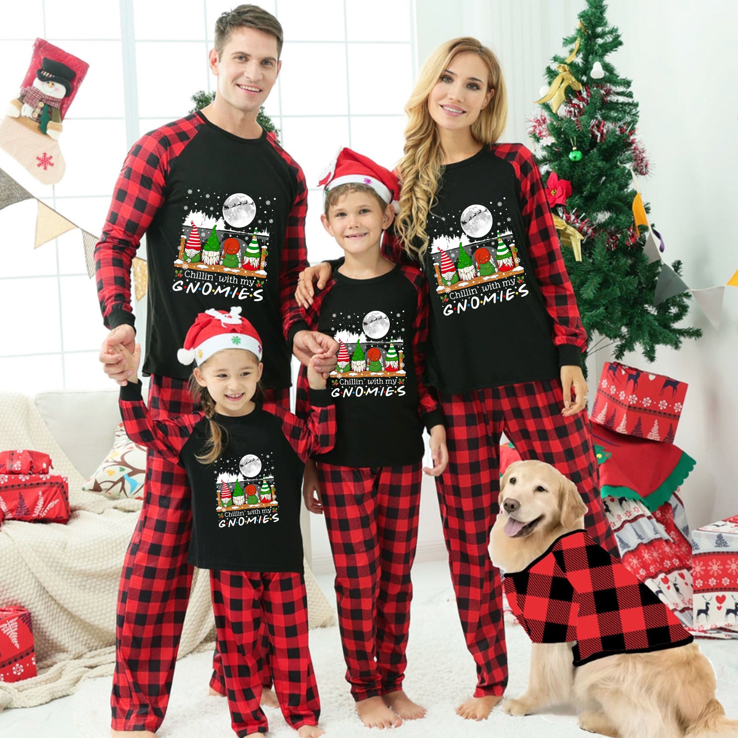 A family of four wearing matching black and red plaid Christmas pajamas with a 'Gnomes' graphic on the front, posed in a living room with a Christmas tree, wrapped gifts, and a golden retriever dog wearing a matching pajama shirt.