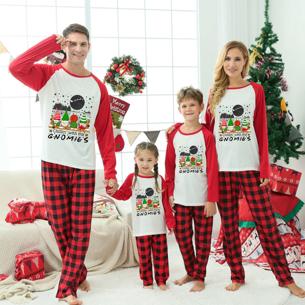 A family of four wearing matching Christmas-themed long-sleeve pajamas stands together in a decorated living room. The pajamas have red and black buffalo plaid legs with white fronts and red sleeves, featuring a festive Santa graphic.