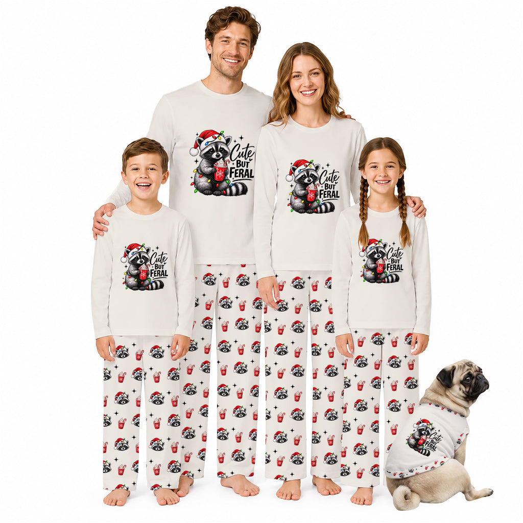 A family of four wearing matching Christmas-themed long-sleeve pajamas and matching pants, standing together on a white background with a small dog wearing a matching pajama shirt at the lower right. The pajamas are off-white with a black and red raccoon graphic and the text 'Cute Feral' on the chest, and the pants have an all-over print of the raccoon graphic.