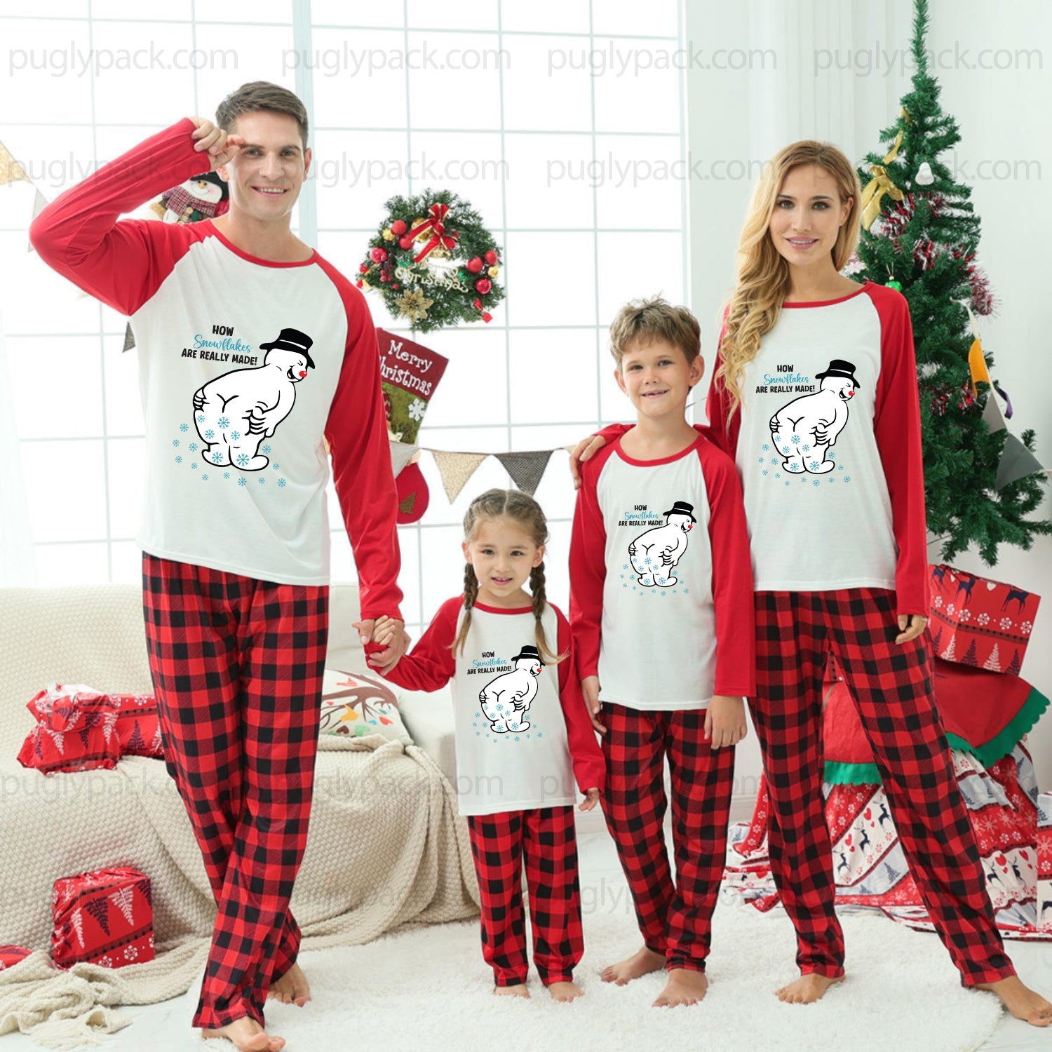 A family of four wearing matching Christmas-themed pajamas, standing together in a decorated living room with a Christmas tree, wreath, and holiday decorations. The pajama set features a White and Red long-sleeve top with a cartoon snowman graphic and Red-and-Black buffalo plaid pants.