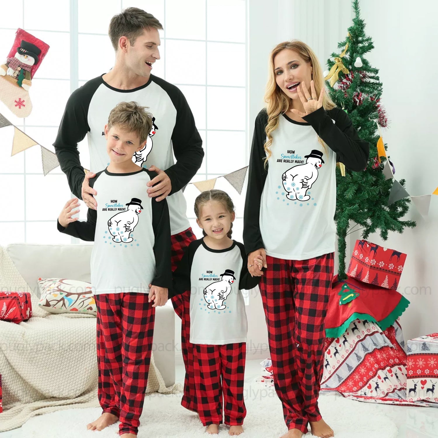 A family of four wearing matching Christmas-themed pajamas, standing in a decorated living room with a Christmas tree, wrapped gifts, and a couch. The matching set features a White long-sleeve top with Black sleeves and a cartoon dog graphic, paiRed with Red and Black buffalo plaid pajama pants.