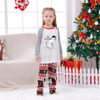A young girl stands in a living room wearing a Christmas-themed raglan long-sleeve pajama shirt and matching patterned pants. The shirt is White on the torso with Gray sleeves and Red collar trim, featuring a cartoon snowman graphic and the text 'Merry Christmas My Dear Frosty'.
