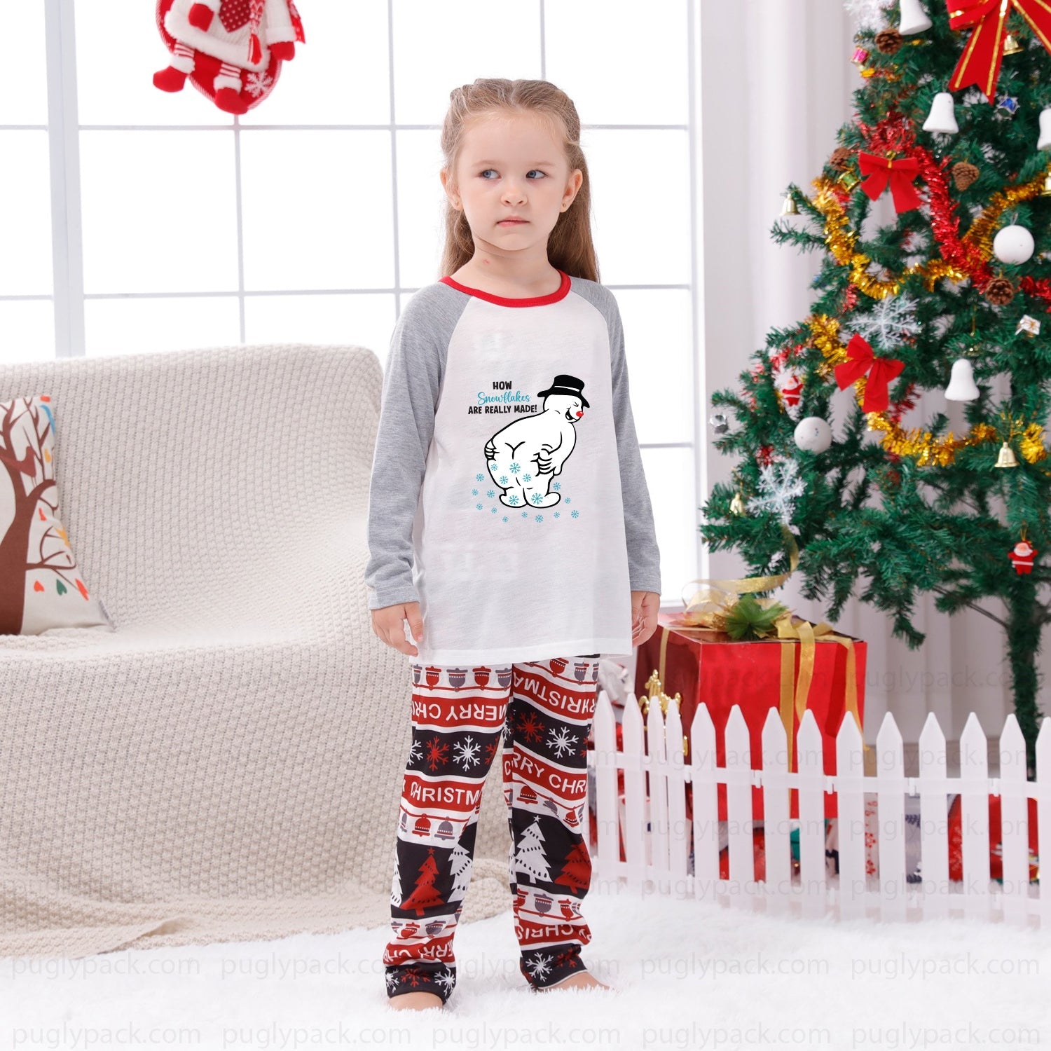 A young girl stands in a living room wearing a Christmas-themed raglan long-sleeve pajama shirt and matching patterned pants. The shirt is White on the torso with Gray sleeves and Red collar trim, featuring a cartoon snowman graphic and the text 'Merry Christmas My Dear Frosty'.