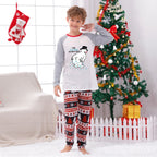A young boy stands barefoot in a decorated living room wearing a Christmas-themed pajama set. The set features a White long-sleeve top with Red raglan sleeves and a cartoon snowman graphic, paiRed with matching Red, White, and Black patterned Christmas pants.