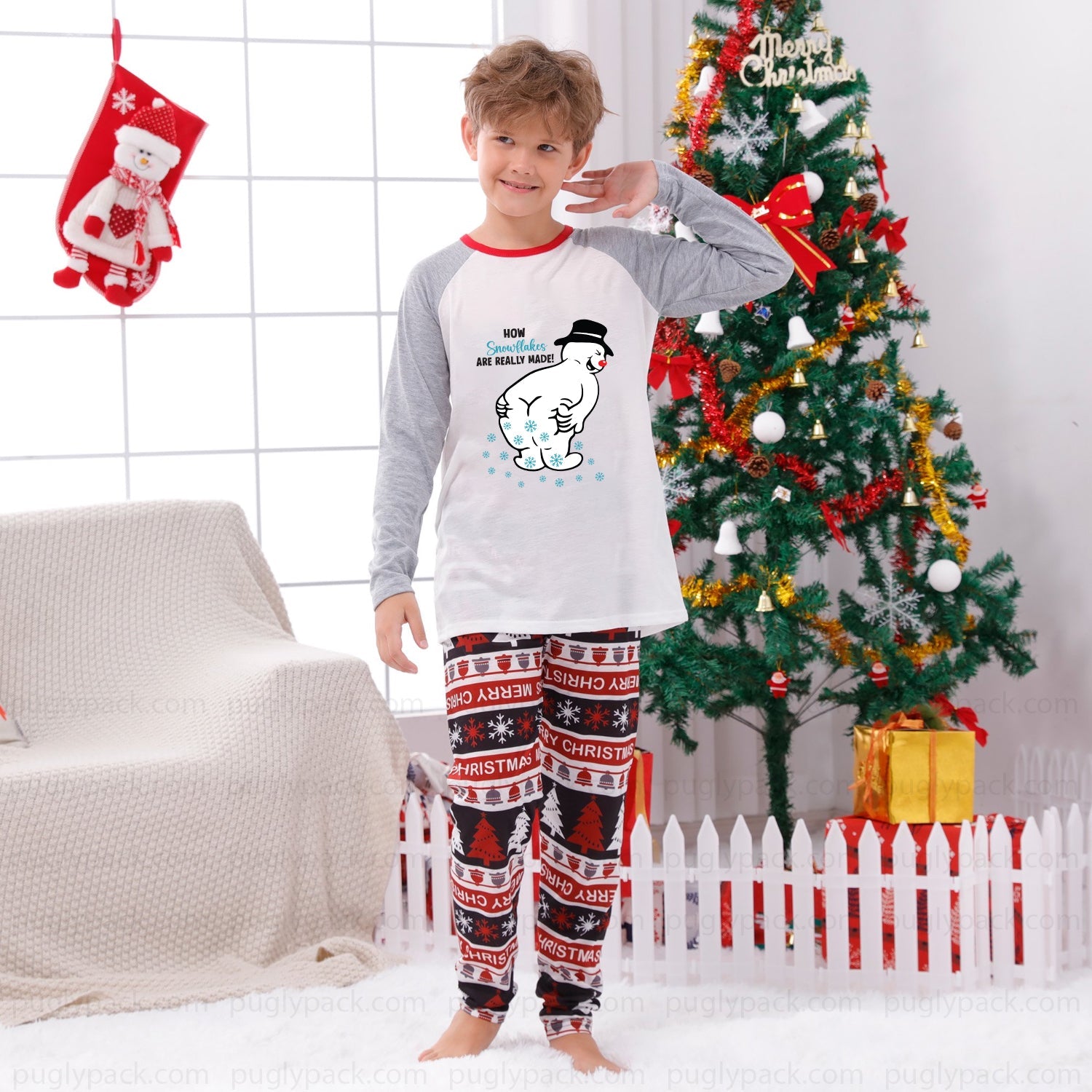 A young boy stands barefoot in a decorated living room wearing a Christmas-themed pajama set. The set features a White long-sleeve top with Red raglan sleeves and a cartoon snowman graphic, paiRed with matching Red, White, and Black patterned Christmas pants.