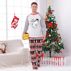 A man modeling a two-piece Christmas pajama set in a decorated living room with a Christmas tree, stocking, and couch. The pajama set features a White long-sleeve top with Red trim and a cartoon polar bear graphic, paiRed with matching Red, White, and Blue patterned pants.