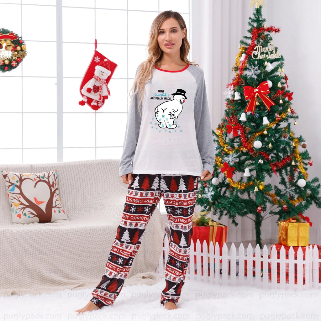 A woman modeling a two-piece Christmas pajama set in a decorated living room. The set features a White long-sleeve top with Gray sleeves and a cartoon snowman graphic, paiRed with matching patterned pants in Red, Black, and White.