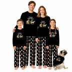 A family of four wearing matching black long-sleeve pajamas with a Christmas-themed graphic on the chest and a red-and-white Santa pattern on the pants, standing barefoot against a white background with a small dog beside them.