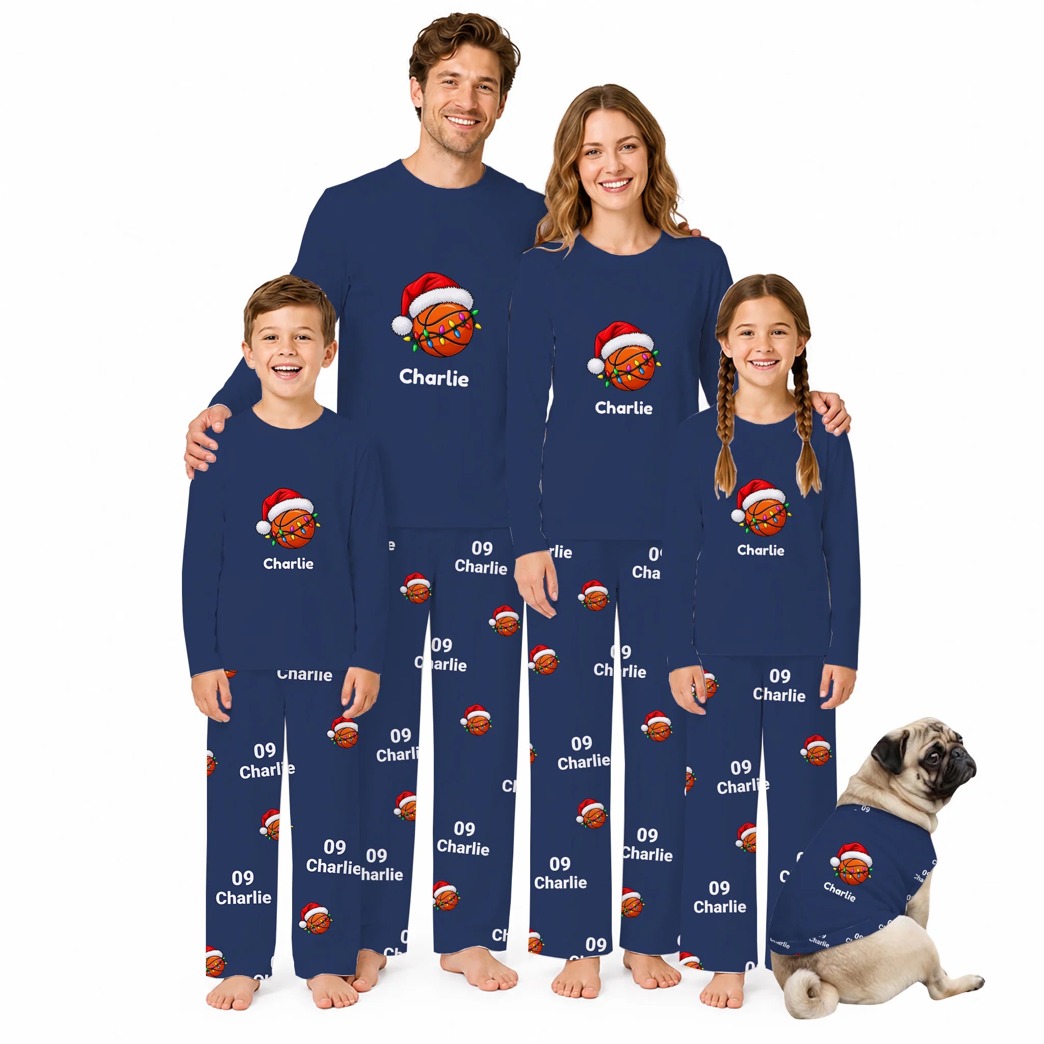 A family of four wearing matching navy blue long-sleeve pajamas with basketball and Santa hat prints, standing together on a white background with a small dog in a matching pajama outfit.