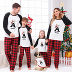 A family of four and their dog wearing matching Christmas pajamas with a white front, black long sleeves, and red plaid pants, standing in a decorated living room with a Christmas tree and stockings.