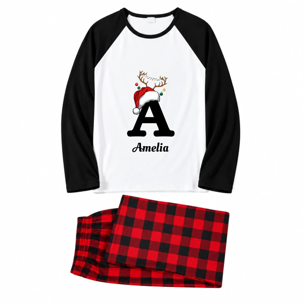 A two-piece pajama set featuring a long-sleeve raglan top with a white body, black sleeves, and a graphic of a black letter 'A' wearing a red Santa hat with reindeer antlers, above the name 'Amelia'. The matching pants are red and black plaid.