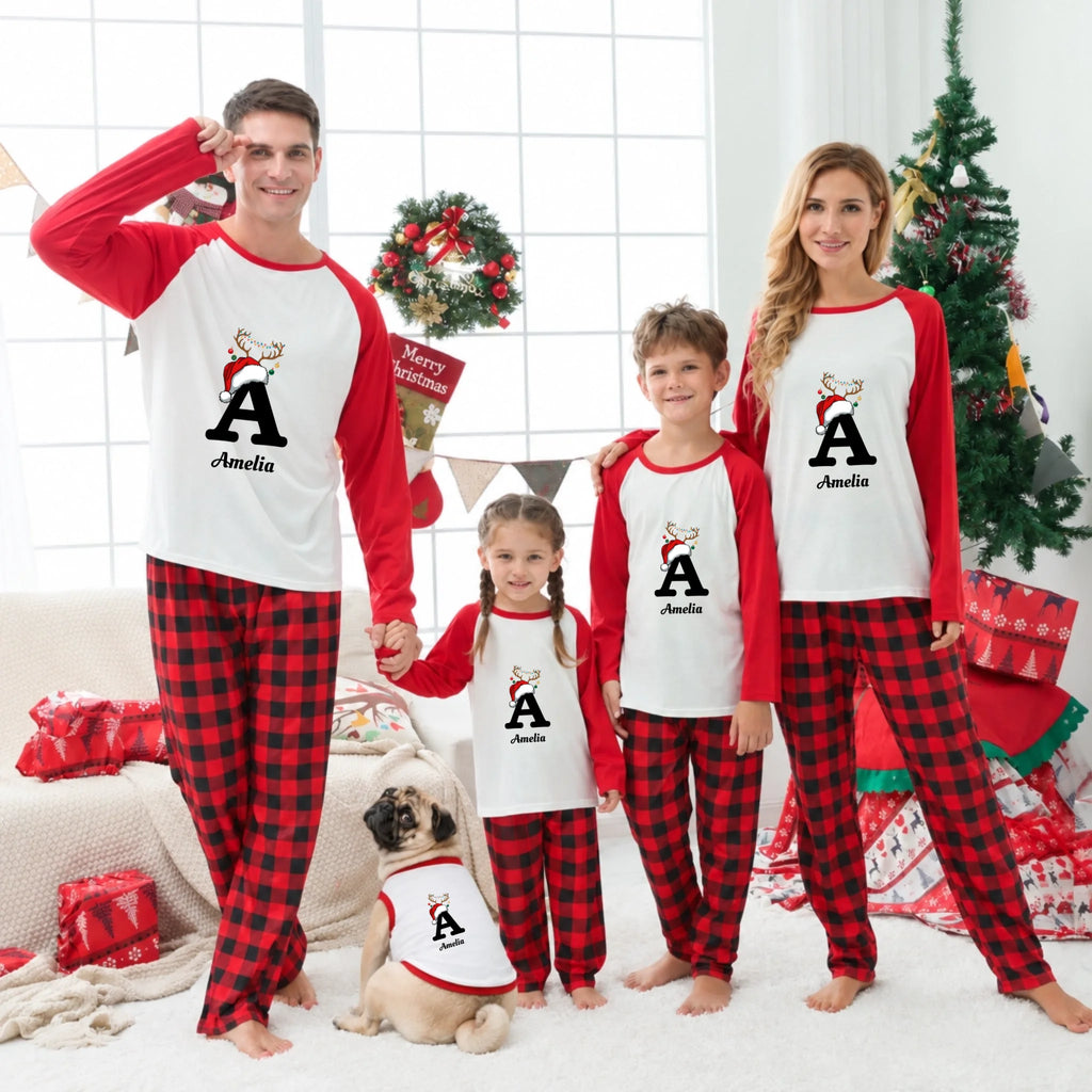 A family of four and a pug dog wearing matching red and black holiday pajamas with white sleeves, standing in a decorated living room with a Christmas tree, gifts, and a window behind them. Each person's pajama top has a large letter A with a reindeer graphic and the name Amelia printed below.